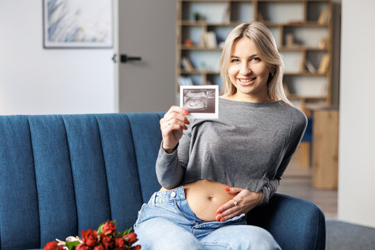 Photo of a pregnant woman at home sitting on couch near beautiful bouquet, holding in her hand baby ultrasound scan print.