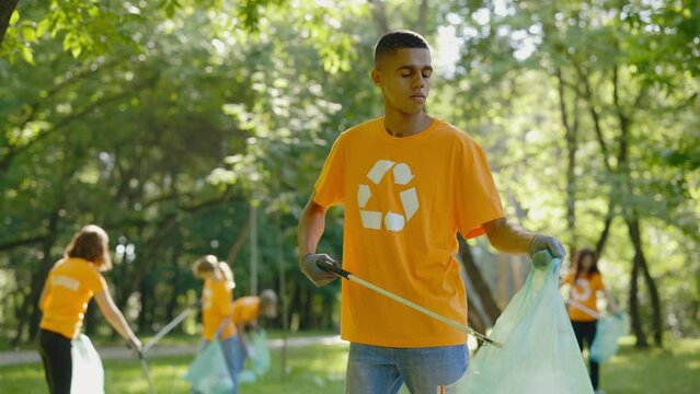 Multiethnic Man Using Trash Picker While Collecting Garbage in Bin Bag. Group of Diverse Volunteers in T-Shirts with Recycling Symbol Cleaning Public Park. Safe Ecology Concept