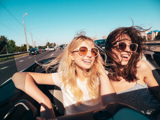 Portrait of two young beautiful and smiling hipster female in convertible car. Sexy carefree women driving. Positive models riding and having fun in sunglasses outdoors. Enjoying summer days - obrazy, fototapety, plakaty
