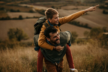 Single father and his son walking in the landscape hills. Piggyback ride boy pointing with a finger...