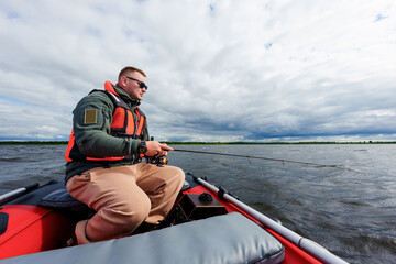 A fisherman is sitting in an inflatable motor boat with a spinning rod. Professional fishing on the lake