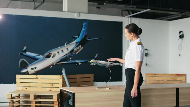 Side view of woman wearing uniform working as pilot instructor. Female pilot walking in classroom, looking, touching airplane model, smiling. Concept of aviation tourism.