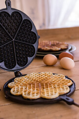 An antique waffle iron with a freshly baked waffle next to it are country eggs and a dish with freshly baked waffles
