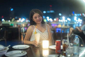 Portrait of an Asian woman holding a cocktail beer alcohol drink glass at night in restaurant on balcony sky bar with urban city skyline background. Sweet fruit food. People lifestyle.