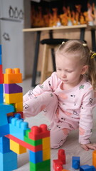 A laughing little child, a blonde preschooler playing with colorful cubes, sitting on the floor in pink pajamas, in a sunny room with a large window at home. Early development of children.