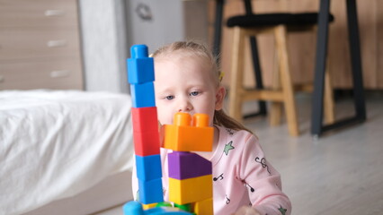 A laughing little child, a blonde preschooler playing with colorful cubes, sitting on the floor in pink pajamas, in a sunny room with a large window at home. Early development of children.