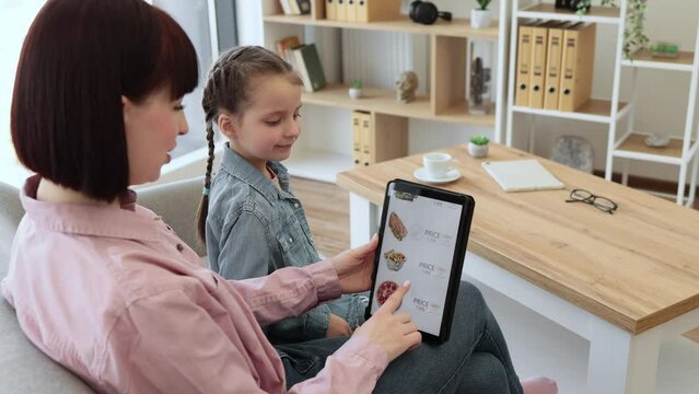 Relaxed mother and daughter searching e-commerce offers on restaurant food with tablet application while having fun indoors. Efficient online clients seizing moment of reduced prices on products.