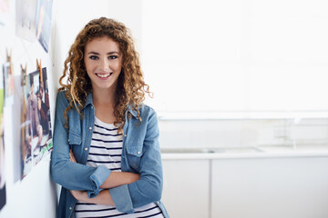 Following a career in graphic design. A young graphic designer smiling at the camera as she stands next to an arrangement of photos on a wall.