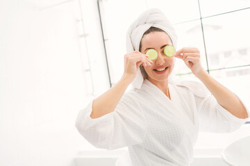 Gorgeous confident adult woman with a towel on her head sitting and putting sliced cucumbers on her eyes in the home bathroom. A pretty attractive woman beauty lifestyle and cosmetic concept.