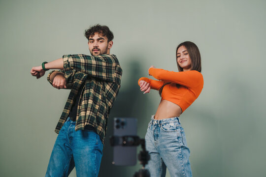 Positive young couple filming video blog for social network, using cell phone on tripod to broadcast their dance isolated on gray wall background. Content