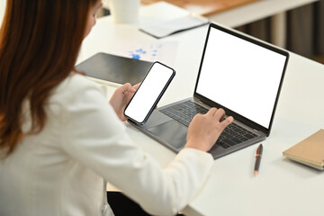 Side view of young female employee sitting at her office desk, hand typing on laptop and using smart phone