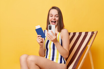 Image of amazed happy young woman wearing striped one-piece swimsuit, holding passport and using...