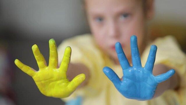 A sad child girl shows her hands painted in the colors of the Ukrainian flag, yellow and blue