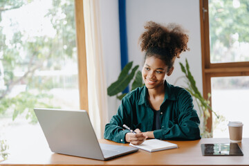 Attractive African woman smiling thinking planning writing in notebook, tablet and laptop working from home,   at office
