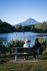 Couple sitting on the bench and enjoying the view of Mt Taranaki at Lake Mangamahoe. New Plymouth. Vertical format.