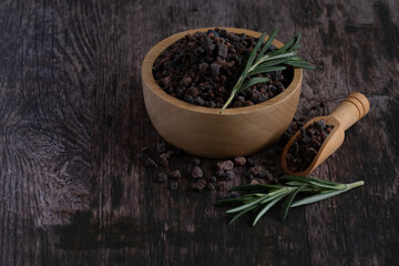 Himalayan black salt, Indian black salt or Kala namak in wooden bowl with wooden scoop and rosemary on wooden table. Used in cooking, especially in Asian and Indian cuisine. Bath salt spa.