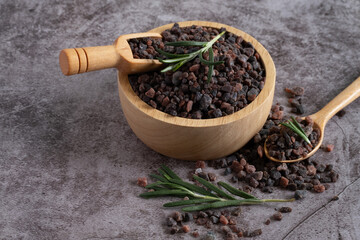 Himalayan black salt, Indian black salt or Kala namak in wooden bowl with wooden scoop and rosemary on black stone table. Used in cooking, especially in Asian and Indian cuisine. Bath salt spa.