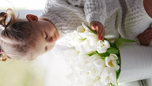 Little girl sitting by window with tulip flowers bouquet. Happy child, indoors. Mother's day, valentine's day or birthday concept.