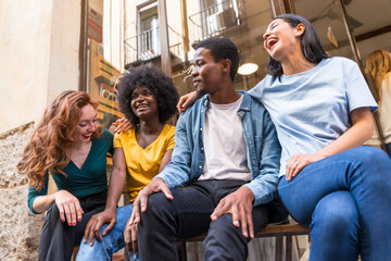 Multi-ethnic friends in a coffee shop sitting in the doorway of the shop window, embracing each other, laughing a lot