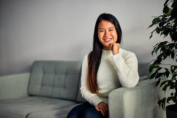Portrait of a smiling Asian adult woman, sitting on a couch, casually dressed.