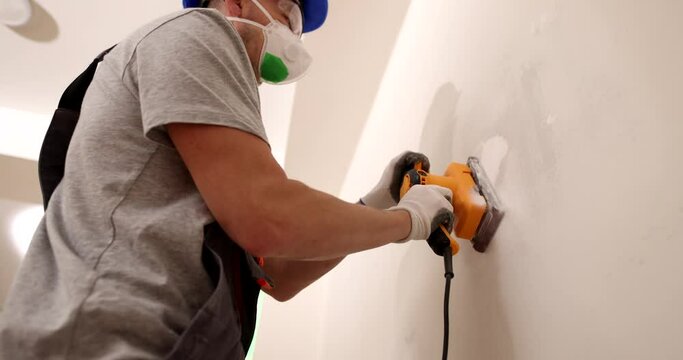 Worker in protective mask and goggles works with grinder to level surface of wall