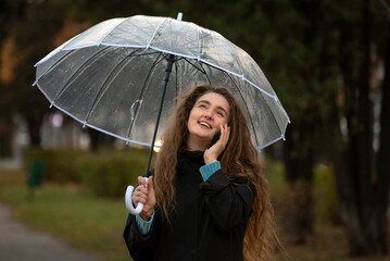 Young beautiful girl in autumn park keeps transparent umbrella talking on phone and laughing.