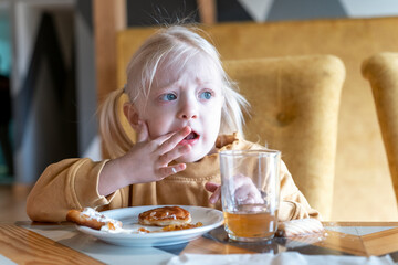 Agitated upset little girl sits at the table and eats dessert pancakes. Cute blonde girl has...