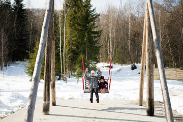 Older brother with younger sister swinging on big wooden swing in forest of mountains.
