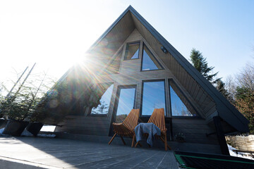 Sunbeam terrace of wooden triangle country tiny cabin house in mountains and two chairs. - obrazy, fototapety, plakaty