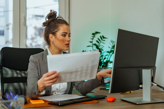 Young woman is sitting at her desk and reading a report on her finance and compares her results on her destop as she gets ready for a meeting with her CEO