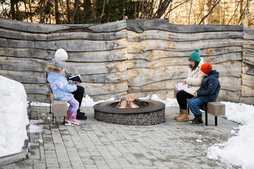 Mother with kids sitting and reading books by camp bonfire on winter in forest. Children in countryside.