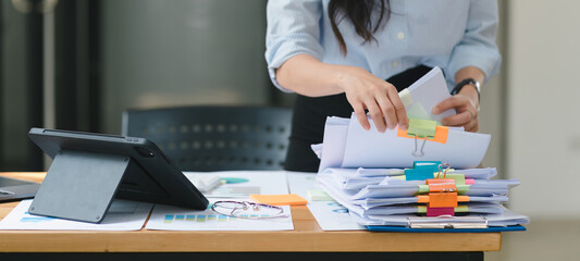A businesswoman is sifting through stacks of paper files and folders that contain both incomplete...