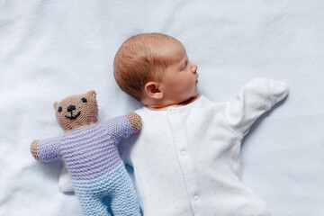 A newborn baby in a white romper is sleeping with soft toys on a white blanket.