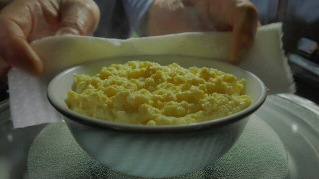Closeup of a man’s hands opening the door of a microwave oven, then wrapping paper towel around a steaming hot bowl of scrambled egg, then lifting it out.