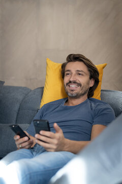 Man watching tv and holding his phone while relaxing on his couch 