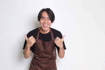 Portrait of attractive Asian barista man in brown apron with thumb pointing away on empty space. Advertising concept. Isolated image on white background