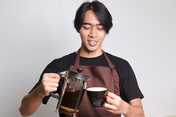 Portrait of attractive Asian barista man in brown apron pouring coffee into a cup from French press coffee maker. Isolated image on white background