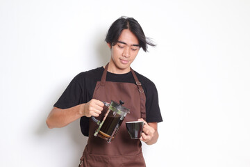 Portrait of attractive Asian barista man in brown apron pouring coffee into a cup from French press...