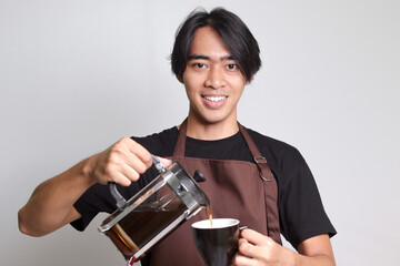 Portrait of attractive Asian barista man in brown apron pouring coffee into a cup from French press coffee maker. Isolated image on white background