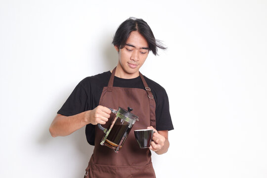 Portrait of attractive Asian barista man in brown apron pouring coffee into a cup from French press coffee maker. Isolated image on white background