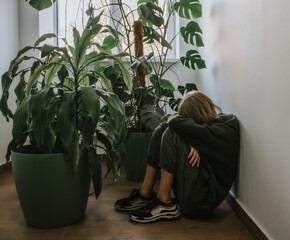 A teenage girl sits with her arms clasped on her knees on the floor against a wall next to houseplants. She's upset and sad. The problem of loneliness, bullying, mental health in the modern world