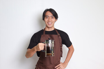 Portrait of attractive Asian barista man in brown apron pointing with finger and showing French press coffee maker. Isolated image on white background