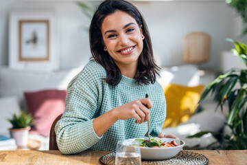 Beautiful smiling woman eating healthy while looking at camera at home. - obrazy, fototapety, plakaty