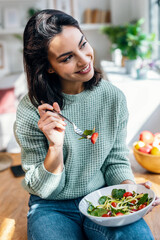 Beautiful smiling woman eating healthy salad while sitting on the table at home.