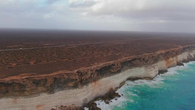 Cinematic drone shot of Campervan on empty road in Nullarbor SA, Australia during cloudy day - Ocean waves crashing against steep rocky coastline