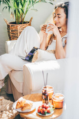 Attractive young woman relaxing in armchair and having breakfast at home.