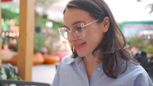 Close up side view young woman face in eyeglasses. Caucasian brunette female wearing glasses use laptop in outdoor cafe. Smiling business woman with spectacles looking computer