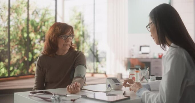 Female Physician Measuring Blood Pressure to a Middle Aged Patient During Consultation in a Health Clinic. Asian Family Doctor Working Behind a Desk in Hospital Office