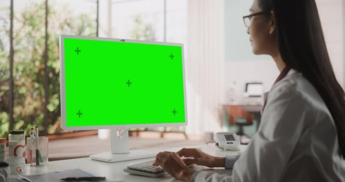 Portrait of an Asian Medical Health Care Professional Working on Desktop Computer with Green Screen Mock Up Display in Hospital Office. Female Clinic Physician Appointing Prescriptions Online