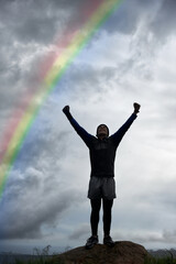 Fototapeta na wymiar Triumph at the top. A young man celebrating at the top of a mountain under a rainbow.